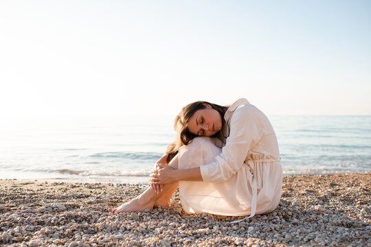 Young woman wearing white linen dress sitting at sea shore with eyes closed over sea. Loneliness