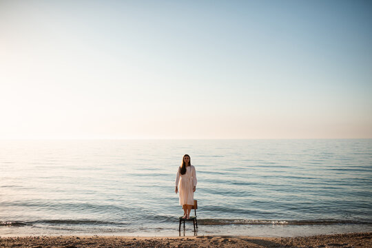 Lonely woman standing on chair in sea water wearing white linen dress at coast line. Loneliness.