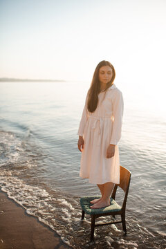Beautiful young woman wearing white linen dress standing on chair in sea water at coastline. Loneliness.