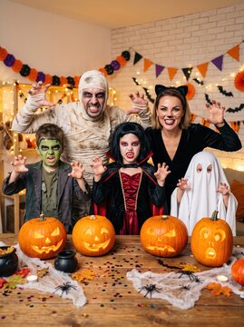 Group of children and adults in scary Halloween costumes posing at festive party table with carved pumpkins and decorations.