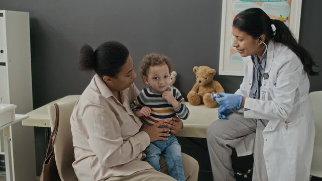 Medium long shot of caring Hispanic female pediatrician examining playful toddler boy sitting on biracial mothers lap in clinic using stethoscope. For pediatric healthcare and family medicine