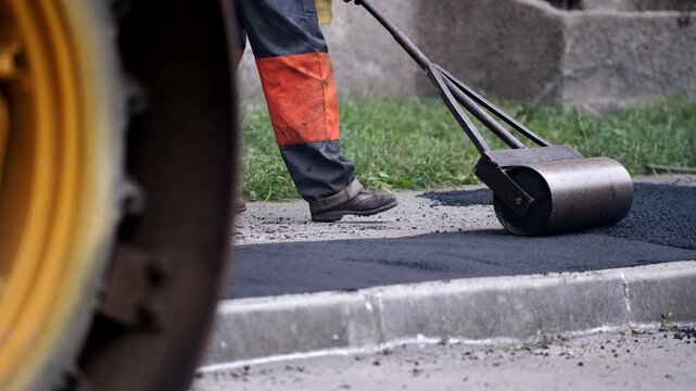 A manual metal roller compacting fresh hot black asphalt on a sidewalk pavement with a yellow industrial vehicle wheel visible in the foreground.