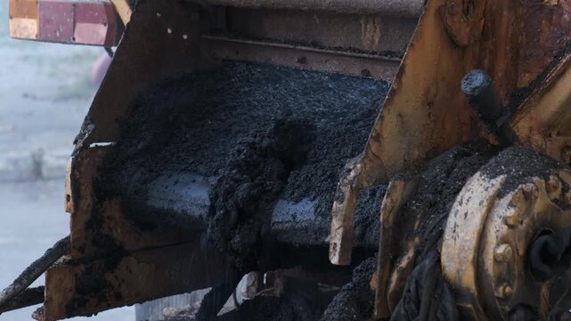A detailed close-up shot of hot, black, steaming asphalt mixture discharging from the hopper of a paving machine during road repair works.