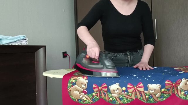 Person from waist up ironing festive teddy bear fabric with red scalloped edges. Holding steam iron over cloth, tidy bedroom with dresser and wardrobe.