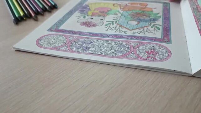 Close up of a hand turning a page in a partially colored adult coloring book. Colored pencils scattered on wooden table, intricate floral and animal illustration visible.