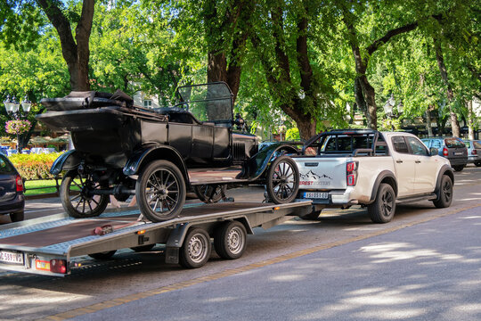 Black Ford Model T (Tin Lizzie) centennial vintage car on a car hauler trailer being towed by a modern Isuzu D-Max pickup past the historic City Hall stone arches in Subotica, Serbia, July 4, 2026
