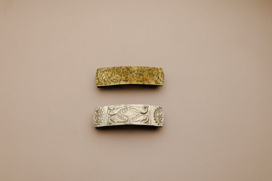 Elegant pair of ornate hair barrettes in metallic gold and silver tones, featuring intricate filigree designs on a subtle beige background