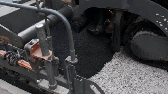 Detailed shot of an industrial paving machine's mechanism distributing hot asphalt mixture onto a crushed stone road base.