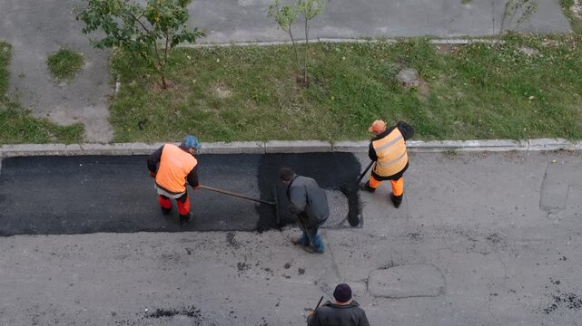 Ukraine, Kiev. June 15 2026. downtown. Top down view of two workers in safety vests spreading and leveling fresh hot asphalt on a city street near a sewer manhole.