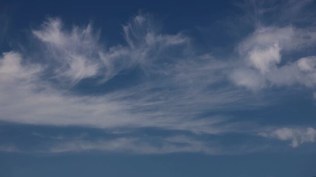 This timelapse footage shows puffy cumulus clouds dissolving into the deep blue sky as thin, wispy cirrus formations gracefully drift across the frame, creating a dynamic atmospheric contrast.