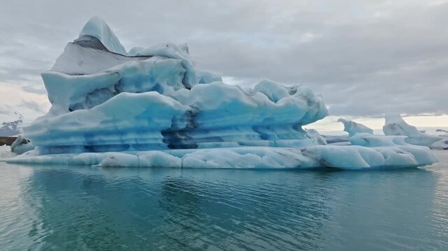 Cinematic aerial side panning around a massive floating iceberg in J&ouml;kuls&aacute;rl&oacute;n Glacier Lagoon, Iceland, showcasing the stunning blue ice, Vatnaj&ouml;kull glacier and Arctic scenery.