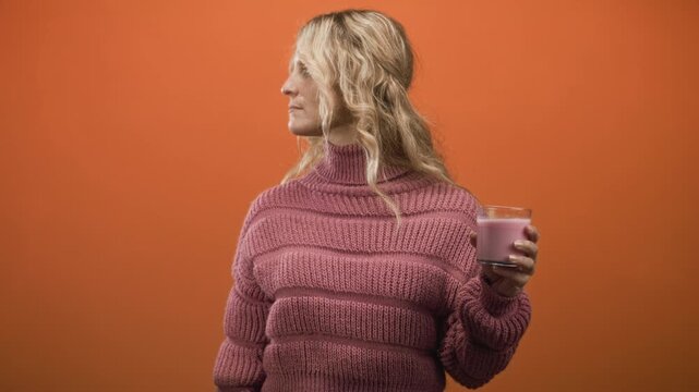 Woman holding strawberry shake glass with hand in studio side profile, wearing pink sweater against orange studio wall; calm reflection.