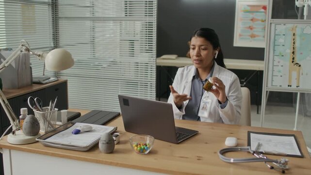 Young Hispanic female doctor wearing lab coat and earbuds talking on video call via laptop, holding pill bottle. Footage is suitable for telemedicine and virtual healthcare marketing