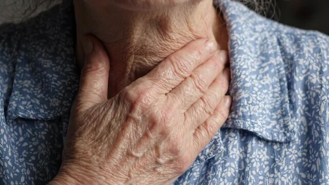 Elderly person rubbing throat with hand showing skin texture wrinkles wearing blue floral shirt expressing discomfort or pain in detail close up view
