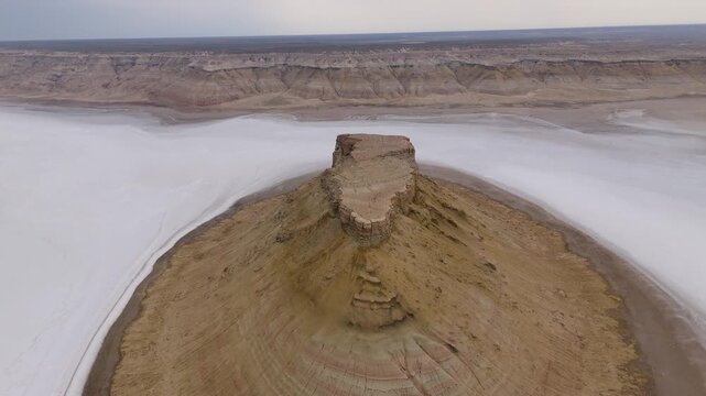 A dramatic close-up drone shot circling a massive rocky outlier monolith rising from the misty, white salt-crusted floor of the Karynzharyk depression in Mangystau, Kazakhstan