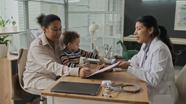 Biracial mother holding toddler signing medical document during consultation with Hispanic female pediatrician in clinic. Perfect for healthcare services and family medical insurance advertisements