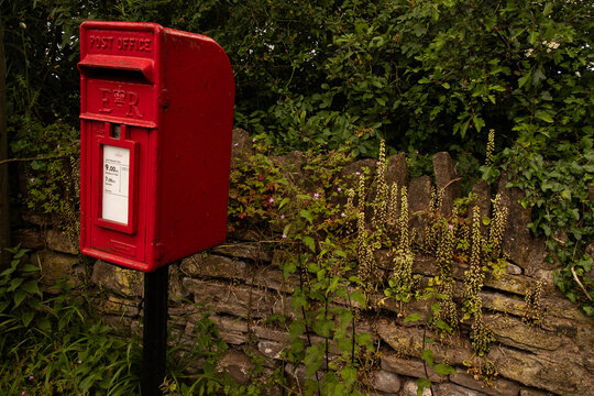 A red British post box on a quiet rural roadside, set against a stone wall, with the royal cipher visible on its front. 