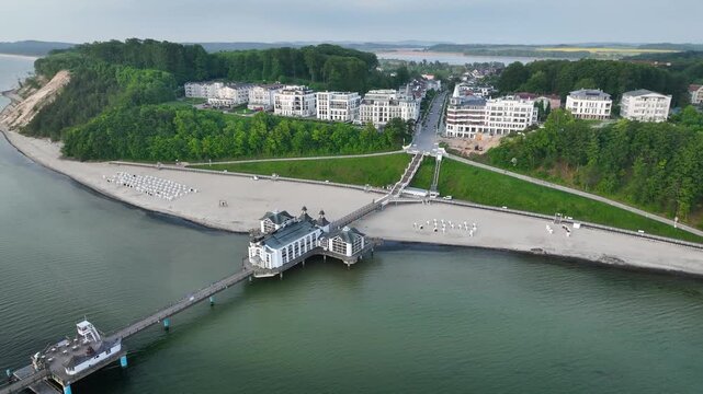 Aerial footage of historic sellin pier and seaside resort on Rugen Island with sandy beach dotted with traditional wicker beach chairs (Strandkoerbe) and  villas along the cliffside.
