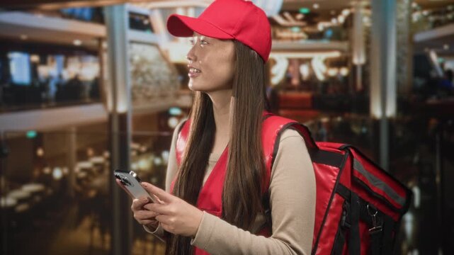 Woman courier holds smartphone and red delivery bag while glancing up in a shopping mall building, visible hands and cap; urban delivery diligence.