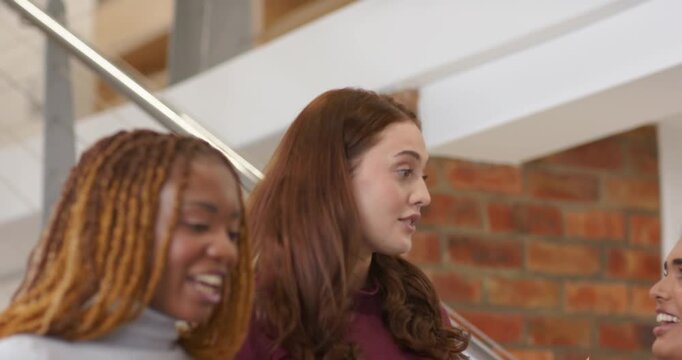 Diverse female friends on metal stairs in lobby, braided woman laughing, central woman explaining