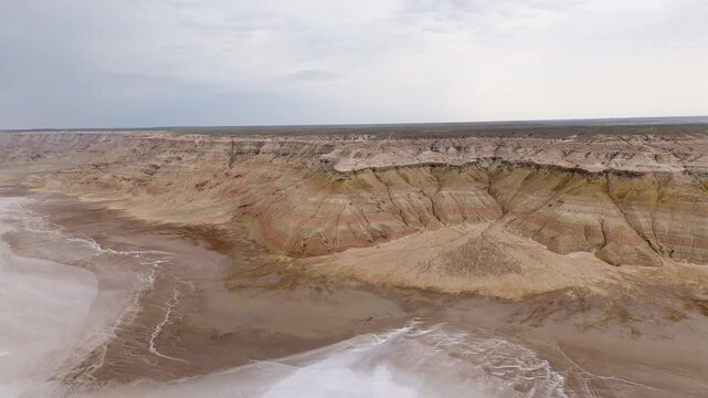 A breathtaking epic drone shot gliding over the misty, water-filled salt flats of the Karynzharyk depression, with spectacular rocky lookout cliffs standing in the foreground within the Mangystau 