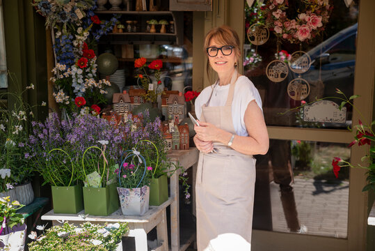 Smiling mature florist holding digital tablet and smiling at the entrance of the flower shop