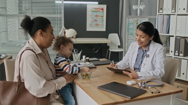 Hispanic female pediatrician consulting with biracial mother holding toddler in clinic. Clip represents pediatric care, family medicine, and diverse healthcare marketing campaigns