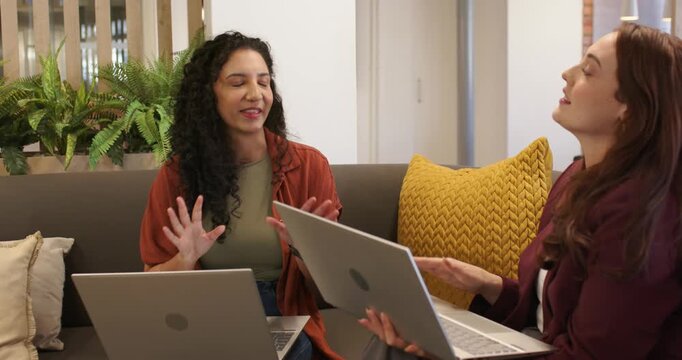 Female colleagues having discussion, left speaker starting talking, gesturing on sofa with laptops