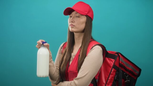 Young woman courier holds milk bottle in hand, wearing red cap and delivery backpack in studio; duty.