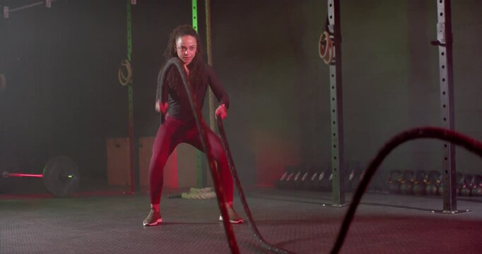 African American woman holding anchored battle ropes producing waves at gym in black top for cardio
