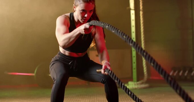 Woman in black gear doing battle ropes in gym, starting pulls for strength, holding them breathing