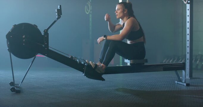 Woman sitting on rower post row wiping forehead, tapping tracker, breathing and resting in gym