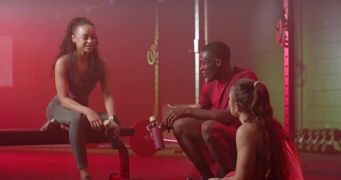 Diverse friends sitting on bench by barbell, passing water bottles and hydrating between sets