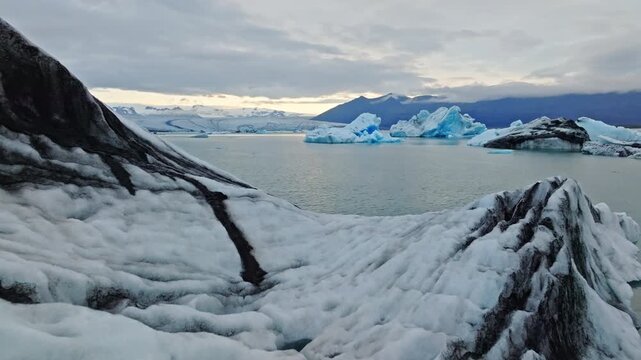 Cinematic aerial flight above floating blue icebergs in J&ouml;kuls&aacute;rl&oacute;n Glacier Lagoon, Iceland, revealing the breathtaking beauty of Vatnaj&ouml;kull glacier and the Arctic landscape at sunset.