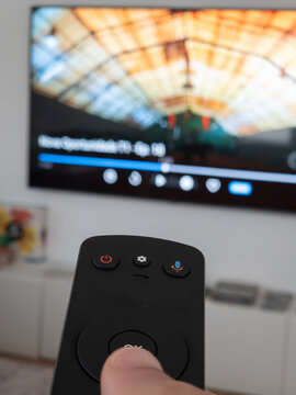 Close-up of a hand holding a TV remote control pointed at a blurred television screen. Concept of home entertainment, streaming, channel selection, media consumption, and modern technology.