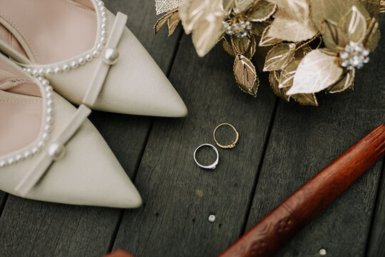 A high-angle composition featuring white bridal heels decorated with pearls, two wedding rings, a traditional golden wedding headpiece, and a wooden Malay keris on a dark wooden floor.