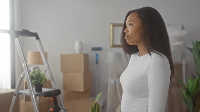 Woman standing in profile showing face looking toward window with moving boxes, ladder and drill in a building; new home hopeful.
