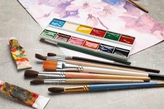 Different brushes, watercolor paints, tubes and drawing on grey table, closeup