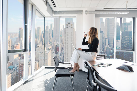 Successful businesswoman working on laptop in modern high-rise office with city skyline view