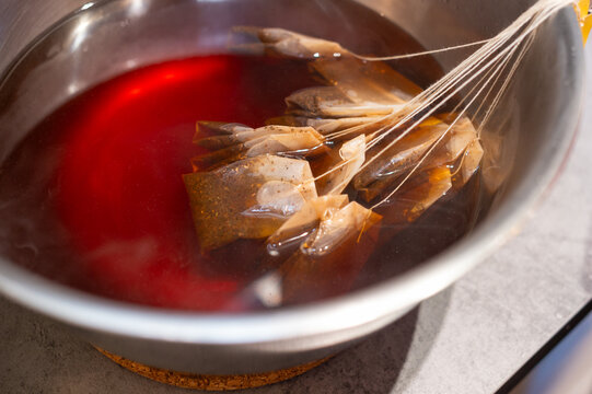 Multiple tea bags are steeping in a large metal bowl filled with red liquid. The scene captures the process of preparing a large batch of iced tea or beverage infusion.