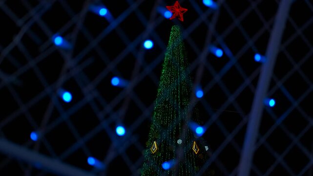 Christmas tree with red star topper seen through decorative lights. Illuminated christmas tree standing tall at night, adorned with vibrant red star and surrounded by twinkling blue decorative lights