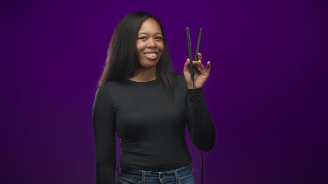 Woman holding a hair straightener in her right hand, smiling and posing with long hair against a purple backdrop in studio; confidence.