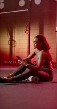 Vertical video: Adult woman sitting on mat with foam roller, scrolling phone after workout in gym