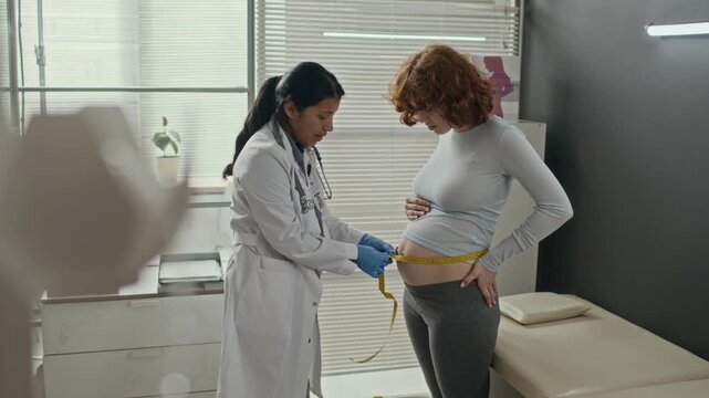 Hispanic female doctor measuring belly of pregnant Caucasian young woman with tape measure in clinic. Perfect for prenatal care, maternal health services, and obstetrics clinic marketing