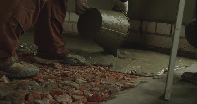 Male worker pouring wet cement mortar from a bucket onto a broken brick floor, preparing the surface for renovation and leveling on a construction site during house repair and remodeling