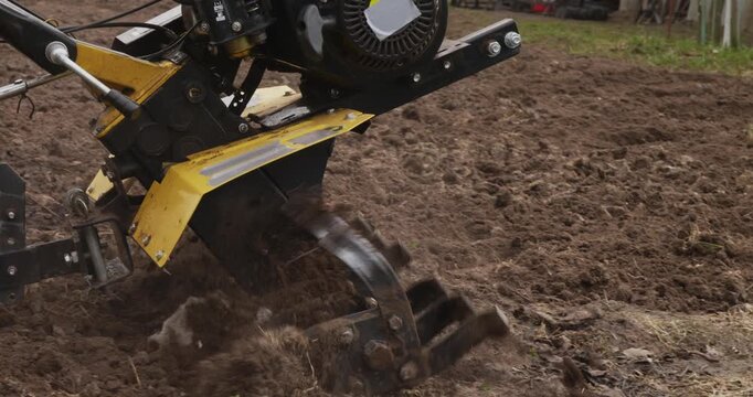 Close-up of a power tiller's rotating blades digging and cultivating the earth, preparing the soil for spring planting and seasonal agricultural work in a backyard garden plot.