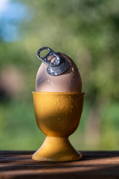 Conceptual egg with aluminum pull tab in holder outdoor, closeup. Creative concept of fresh egg with an easy open metal can tab in yellow ceramic cup on wooden table