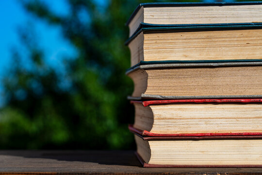 Stack of vintage books on wooden table outdoor, closeup. Stack of old hardcover books on wooden surface against blurry green nature and blue sky background