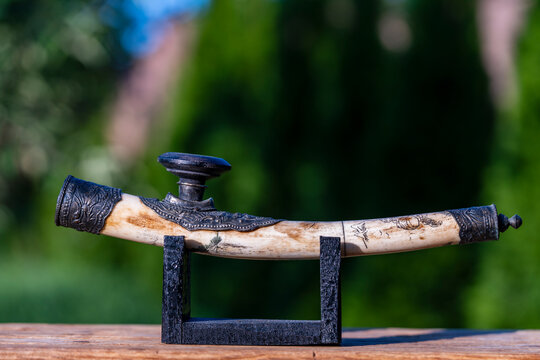 Antique Burmese opium smoking pipe on wooden stand outdoor, closeup. Vintage carved bone opium pipe from Burma with ornate metal details displayed on black wooden stand