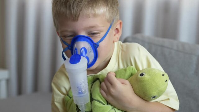 Boy holds green soft toy while breathing through nebulizer mask during home inhalation therapy. Child distraction and comfort during respiratory treatment concept.
